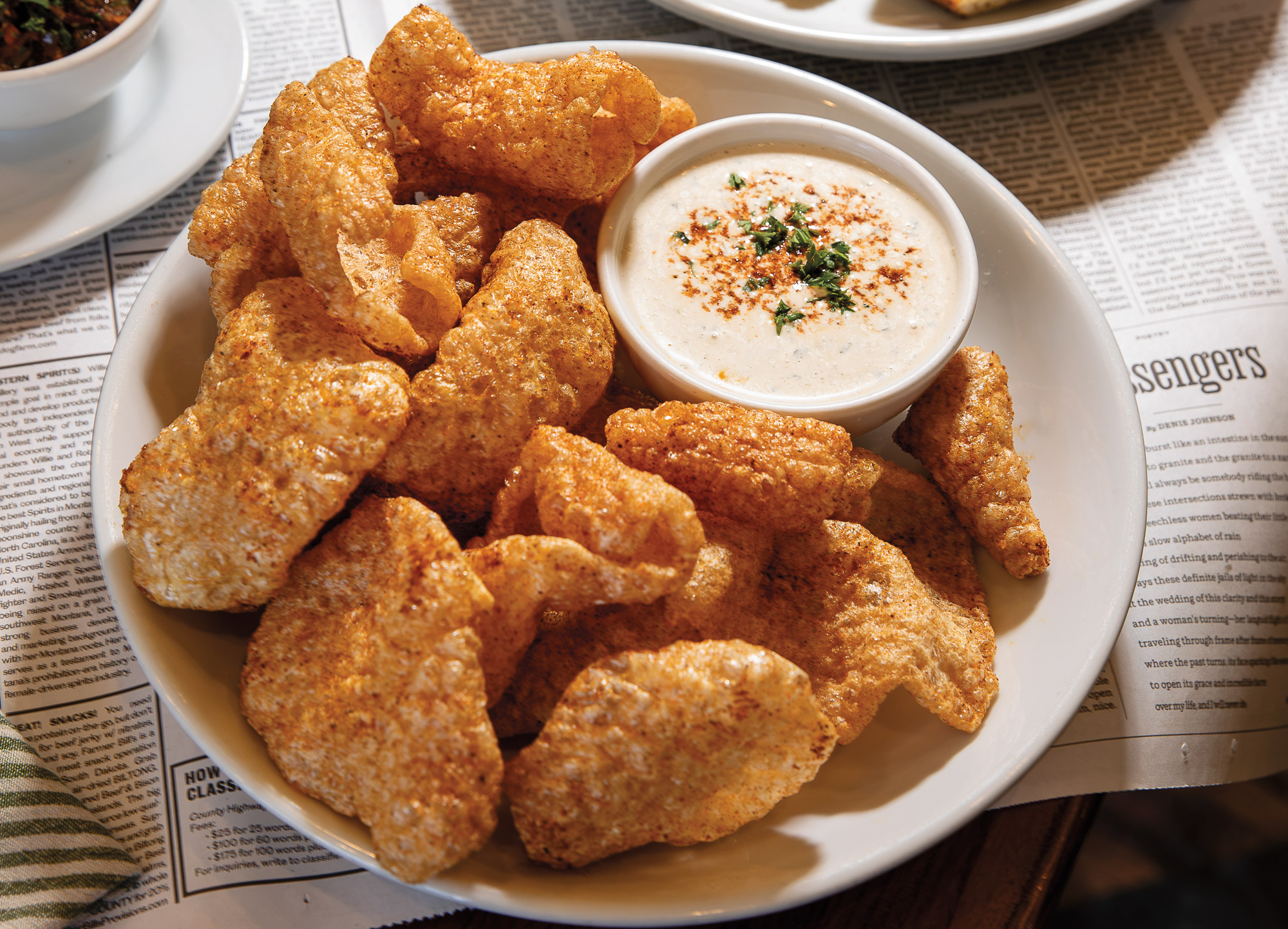 A white plate filled with cracklins and a dipping sauce in a smaller bowl filled with herbs and spices