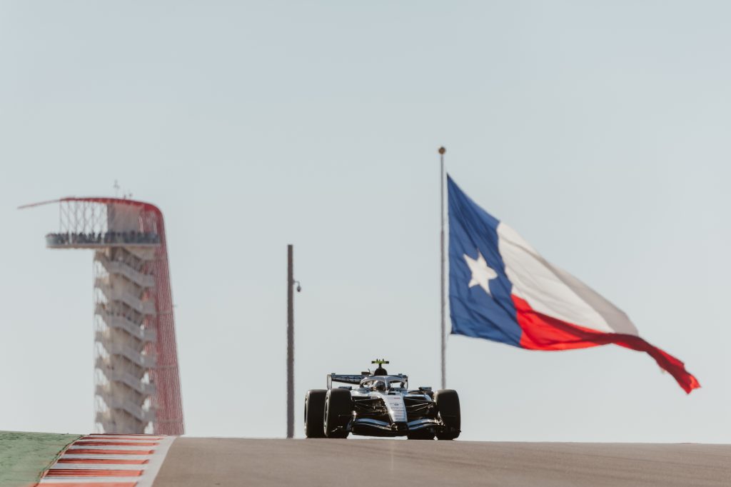 A race car on a track between a large Texas flag and a red and white tower