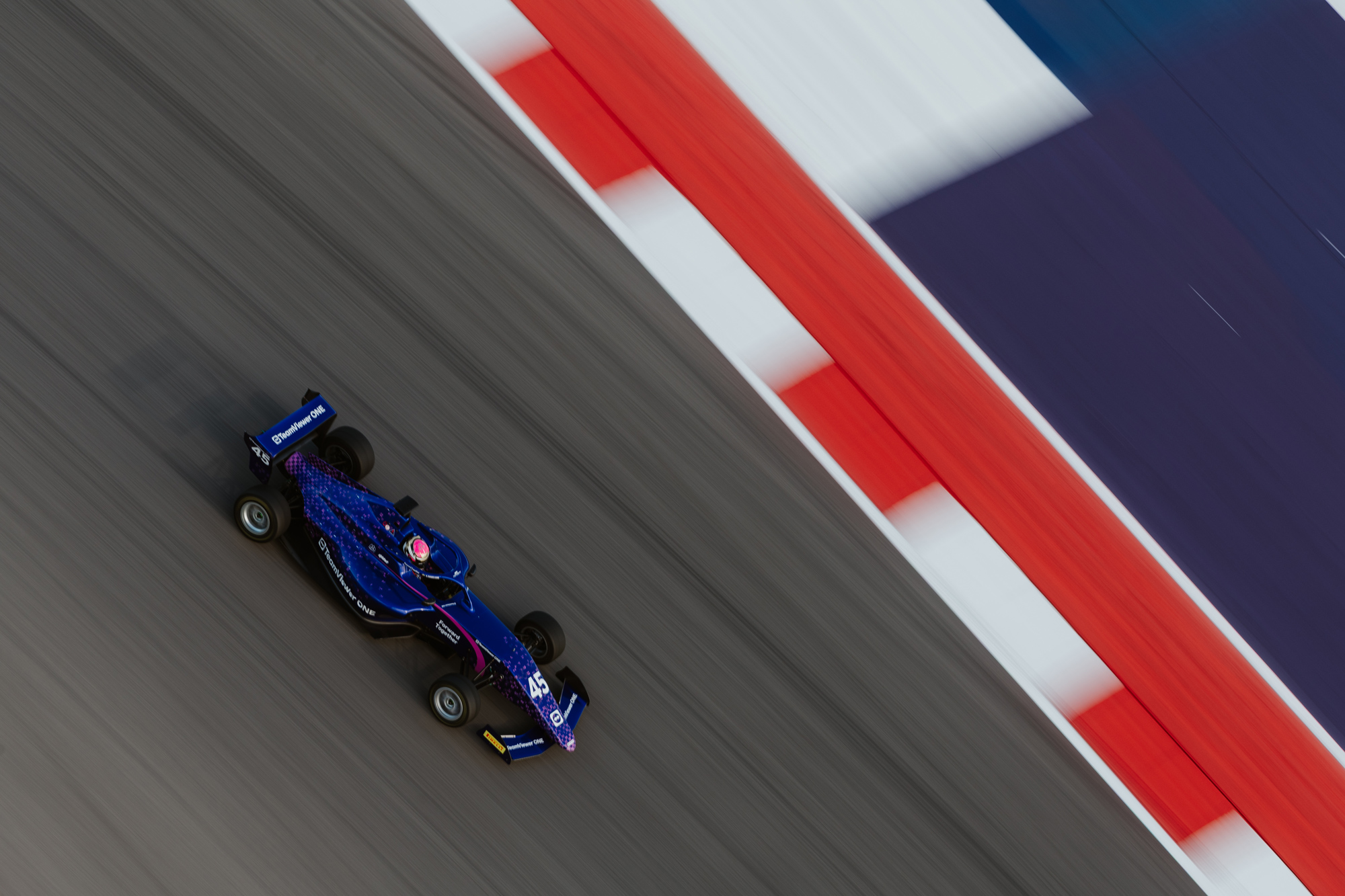 A blue race car driving on a race track with red, white, and blue lines on it