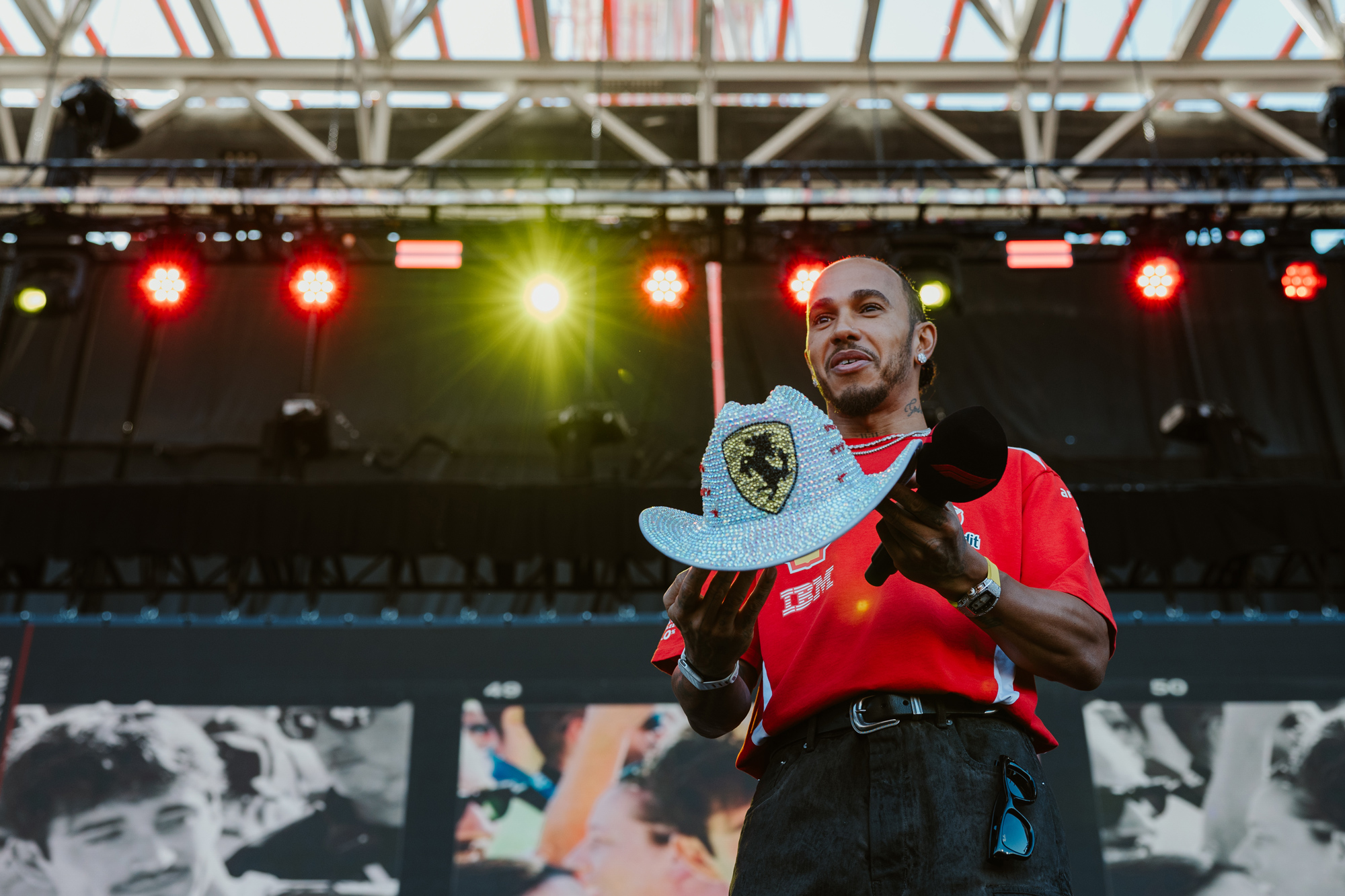 A man in a red shirt and black slacks holds a bejeweled Ferrari cowboy hat and a microphone on a stage