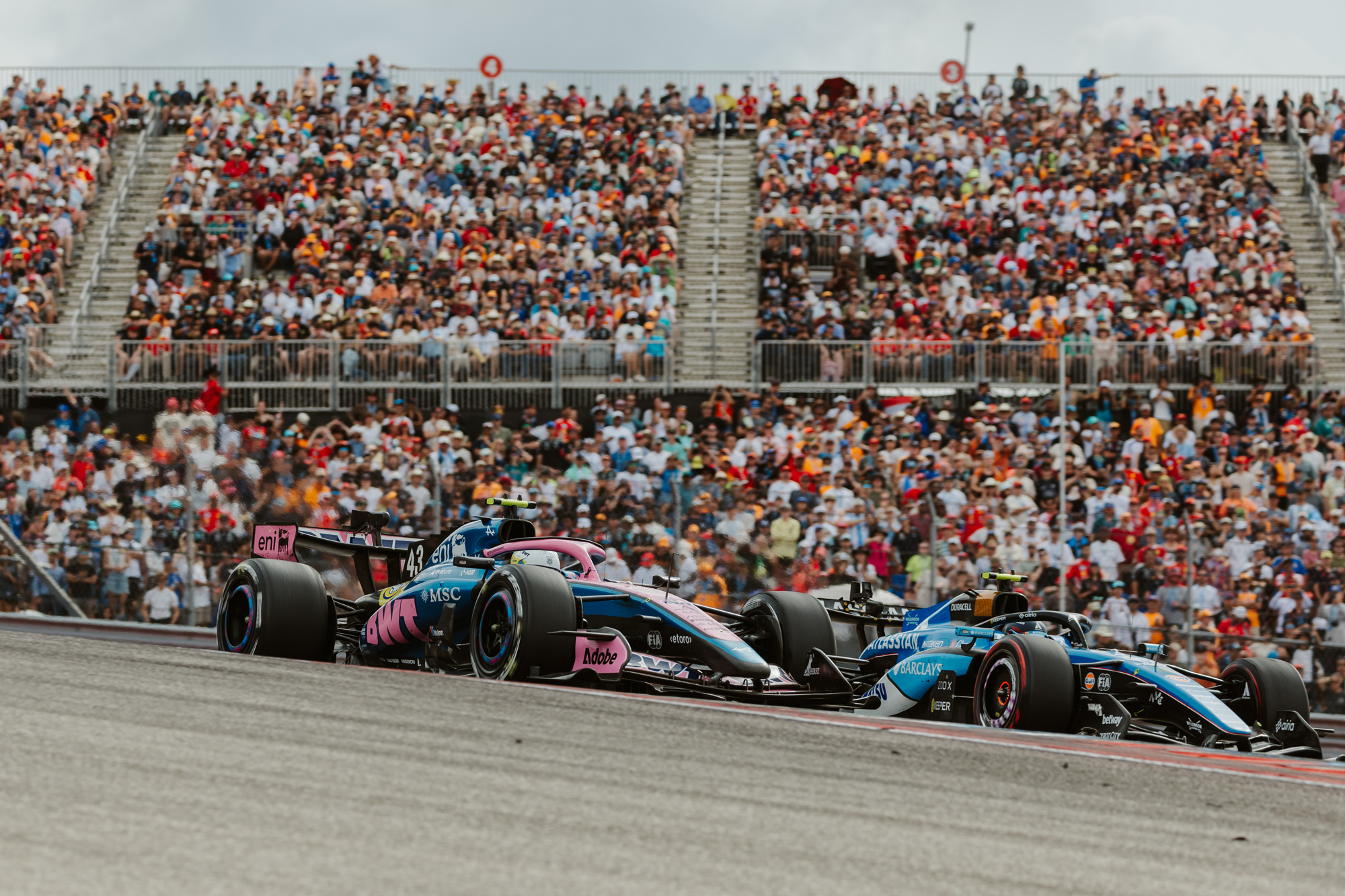 A blue and pink and a blue and white race car next to each other on a track with hundreds of fans behind them in the bleachers