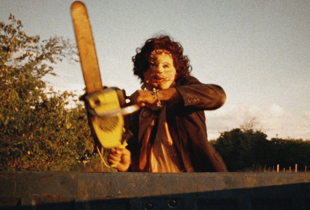A man with a curly wig and a scary mask in a suit and tie wielding a chainsaw