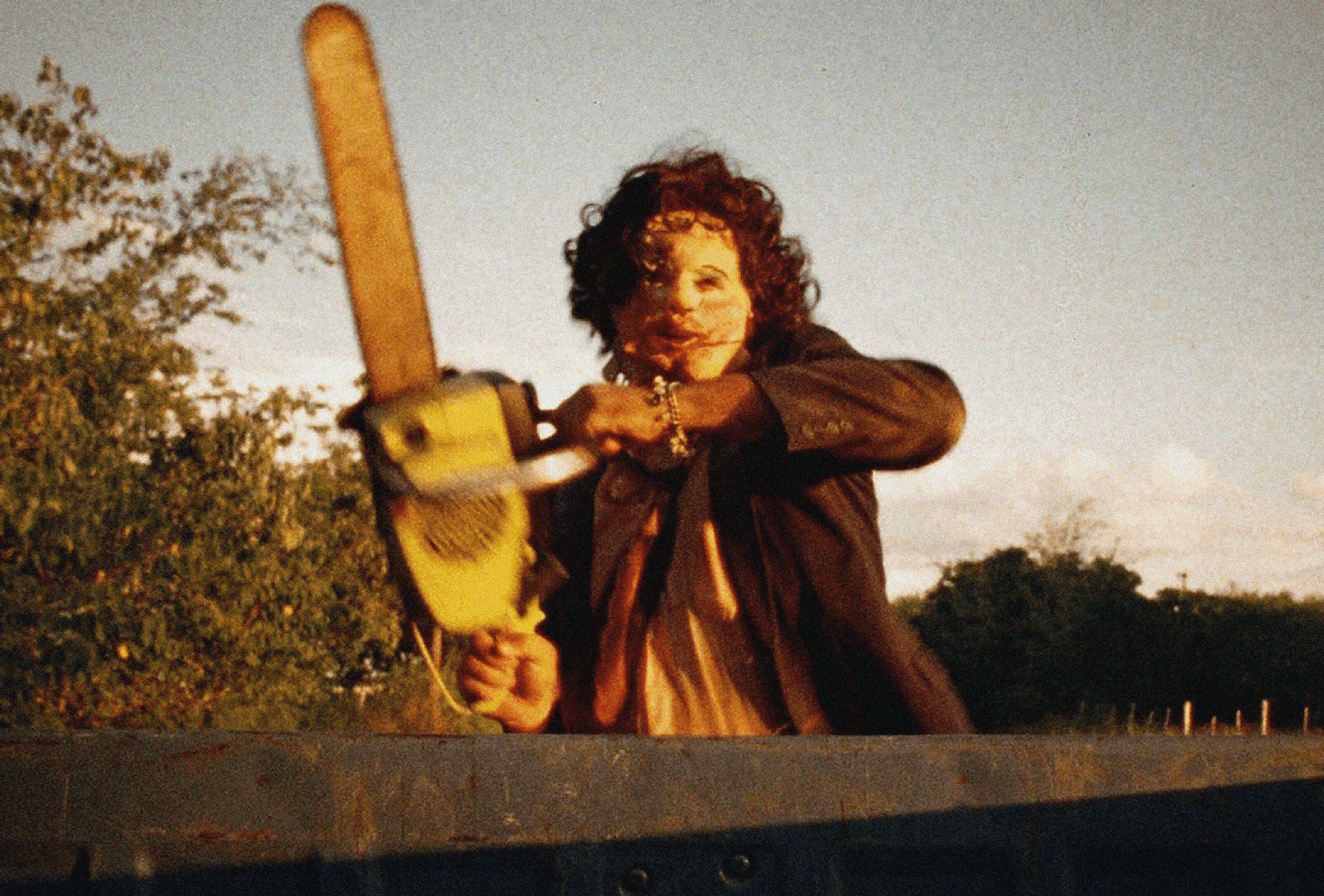 A man with a curly wig and a scary mask in a suit and tie wielding a chainsaw