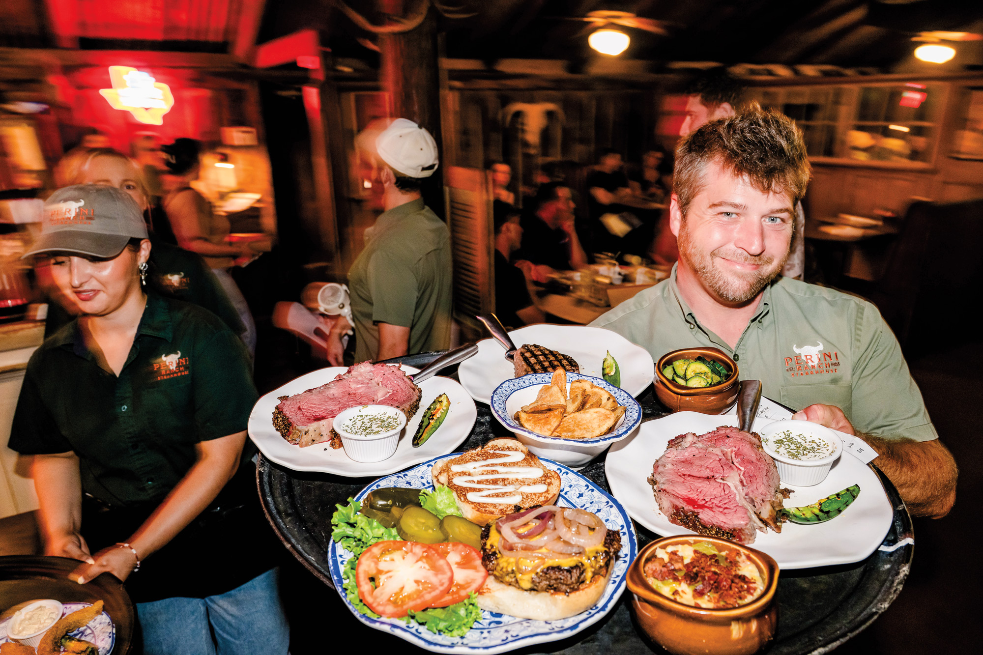 A server with dirty blond hair holds a large tray of food on plates, including a cheeseburger, prime rib, a steak, and a ramekin of queso