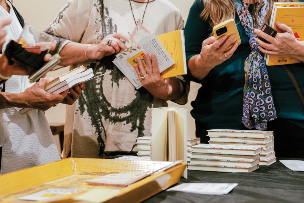 People holding multiple books in their hands, including a person with a colorful scarf and long hair and another in a tie-dyed shirt and multiple rings on their fingers