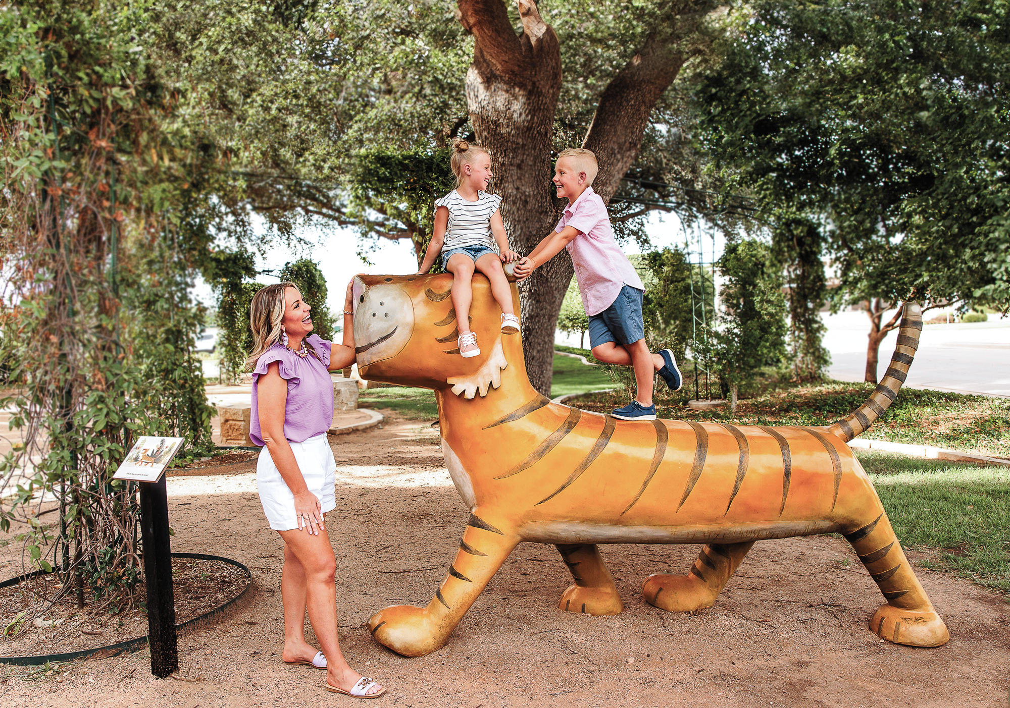Two children atop a statue of an orange tiger with black stripes as a woman smiles and looks on