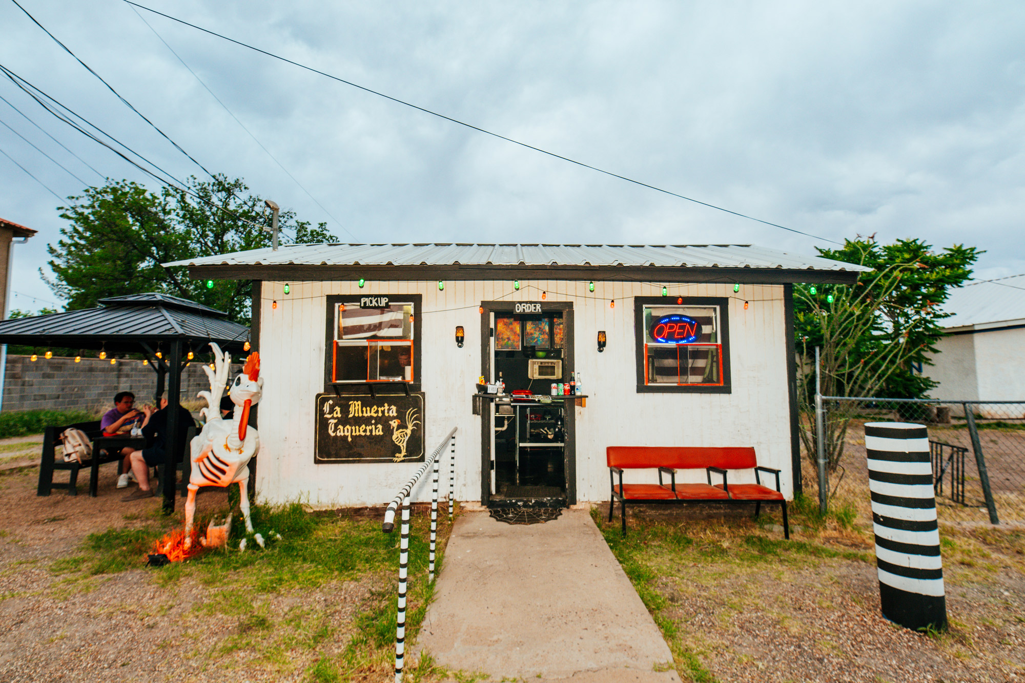 A small white building with a sign that reads La Muerta Taqueria and black-and-white painted features outside including a handrail and a post