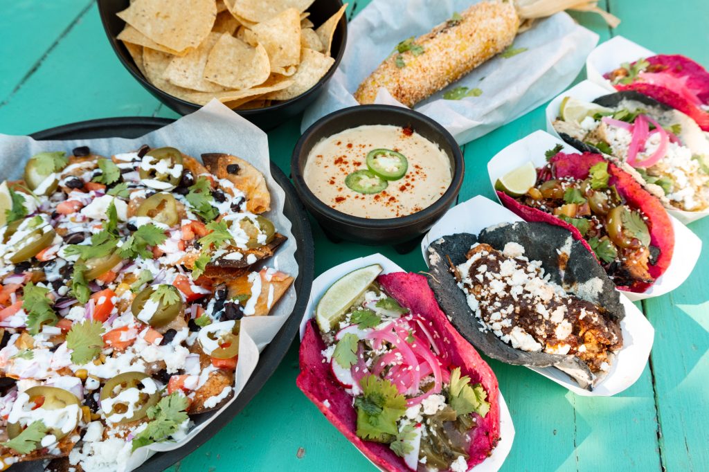 A teal picnic table filled with plates and baskets of food, including tacos with black and pink tortillas, nachos, queso, and tortilla chips.