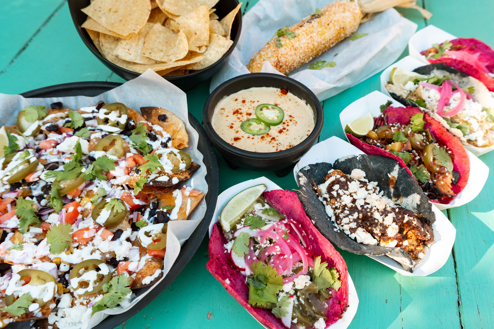 A teal picnic table filled with plates and baskets of food, including tacos with black and pink tortillas, nachos, queso, and tortilla chips.