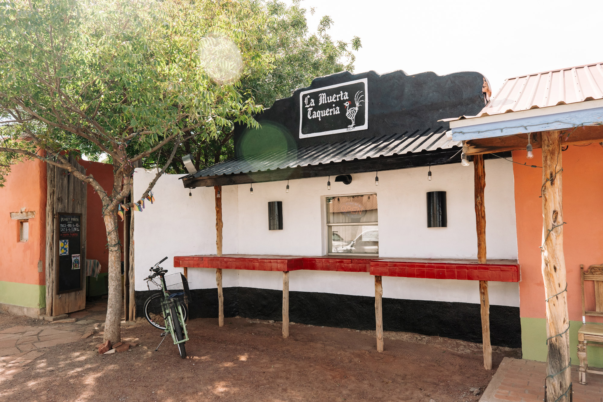 A black, red, and white building with a window in the middle bearing a neon sign that reads OPEN underneath a painted sign that reads La Muerta Taqueria.