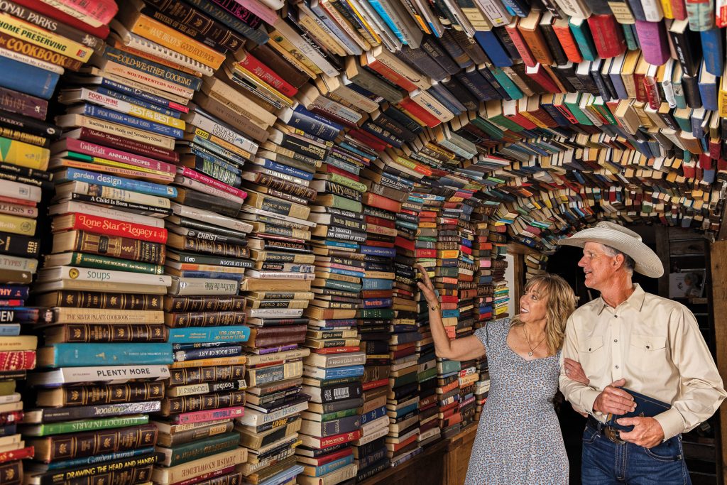 Two people, one in a gray dress, and one in western wear including a cowboy hat, walk under a large archway made of books