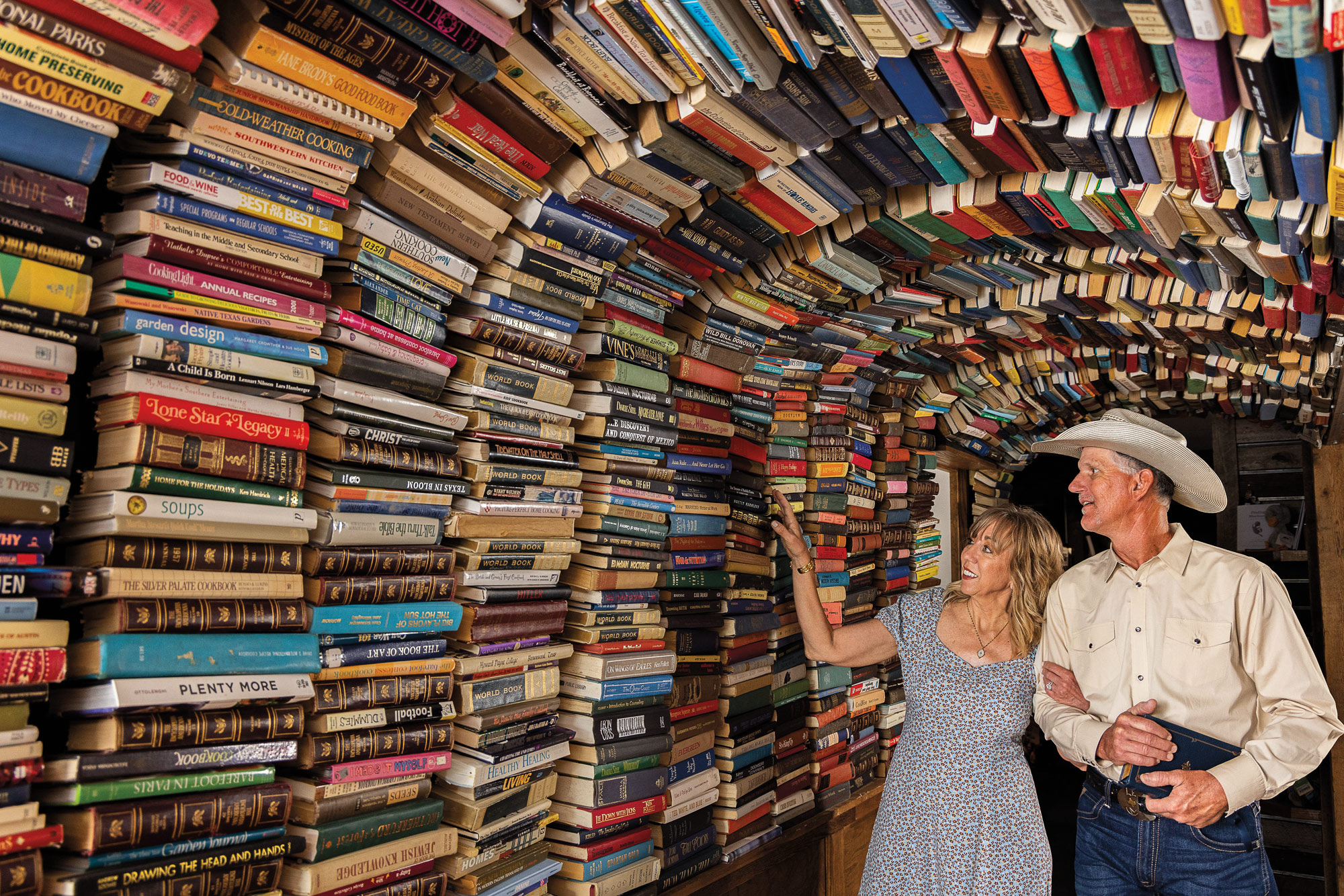 Two people, one in a gray dress, and one in western wear including a cowboy hat, walk under a large archway made of books