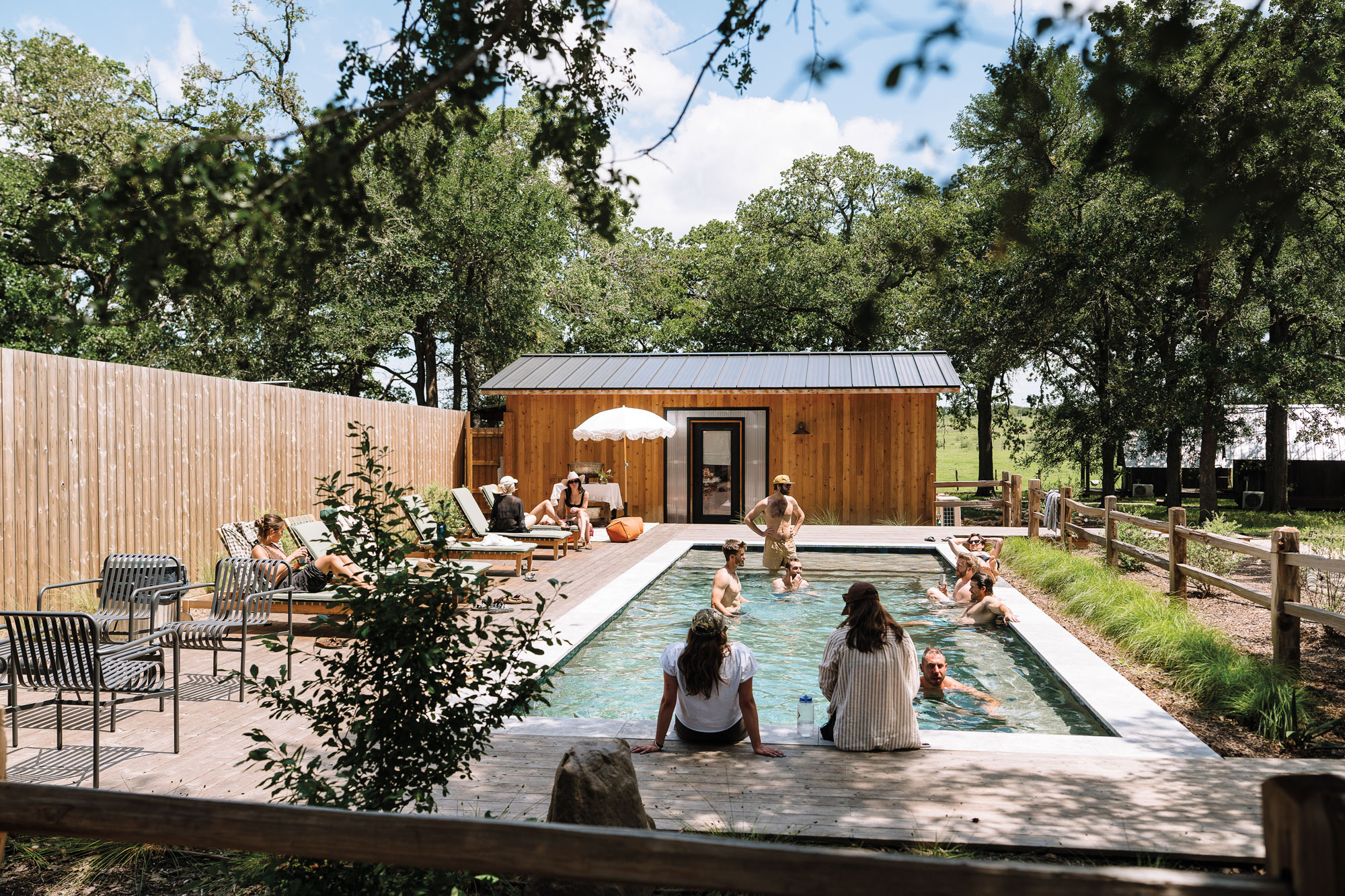 Many people in a rectangular swimming pool and lounging poolside behind a wooden building.