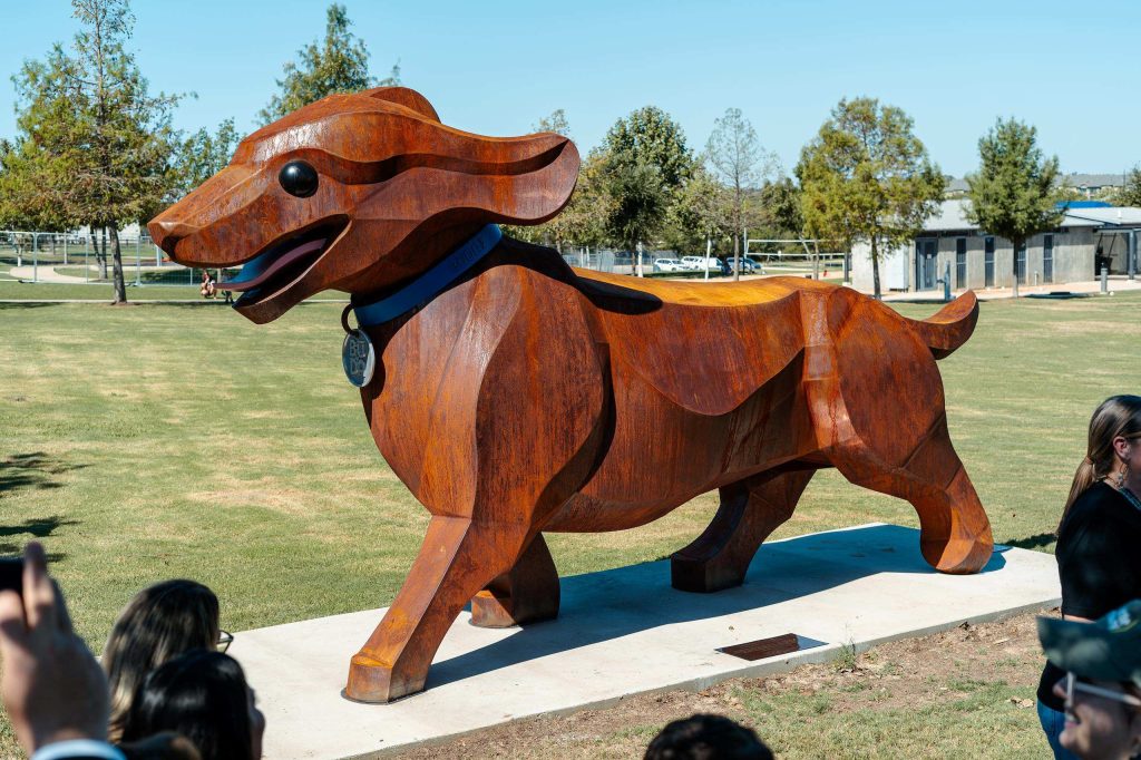 A large wooden statue of a smiling wiener dog wearing a blue collar with trees in the background