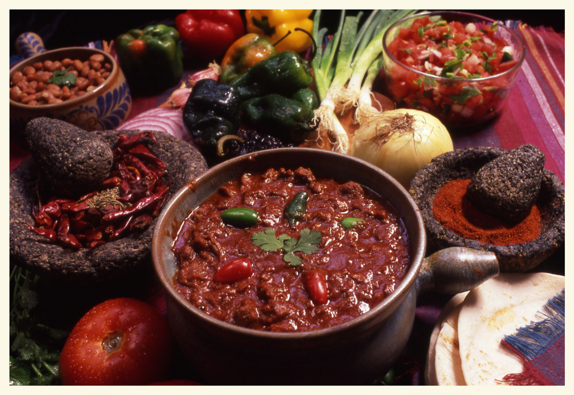 A bowl of chili with red and green peppers in it, surrounded by bowls of condiments like beans, diced tomatoes, ground chili powder, and dried red chilies.