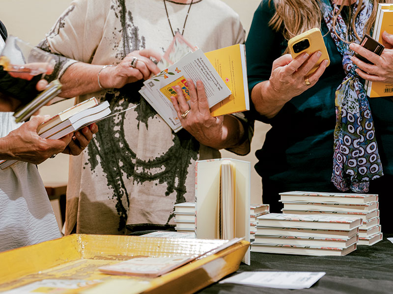 People holding multiple books in their hands, including a person with a colorful scarf and long hair and another in a tie-dyed shirt and multiple rings on their fingers