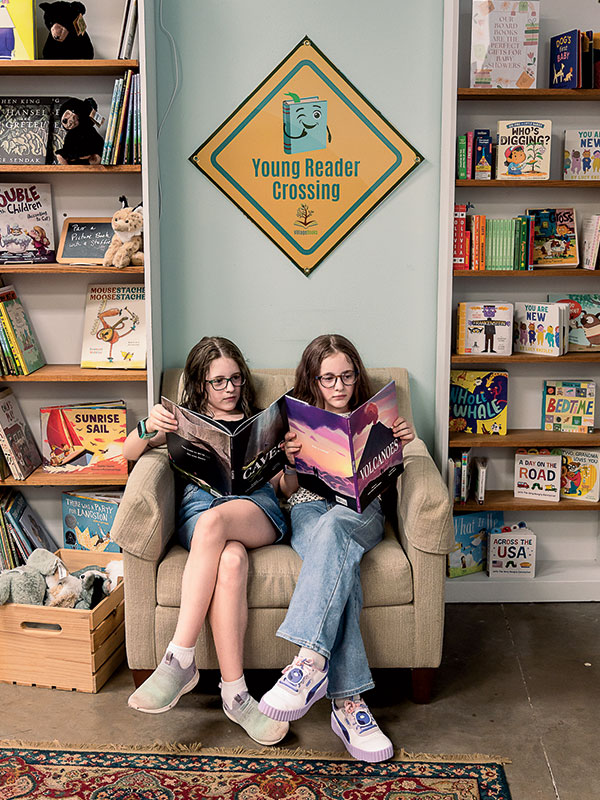 Two young girls sitting cross-legged on a beige chair, each reading a book.