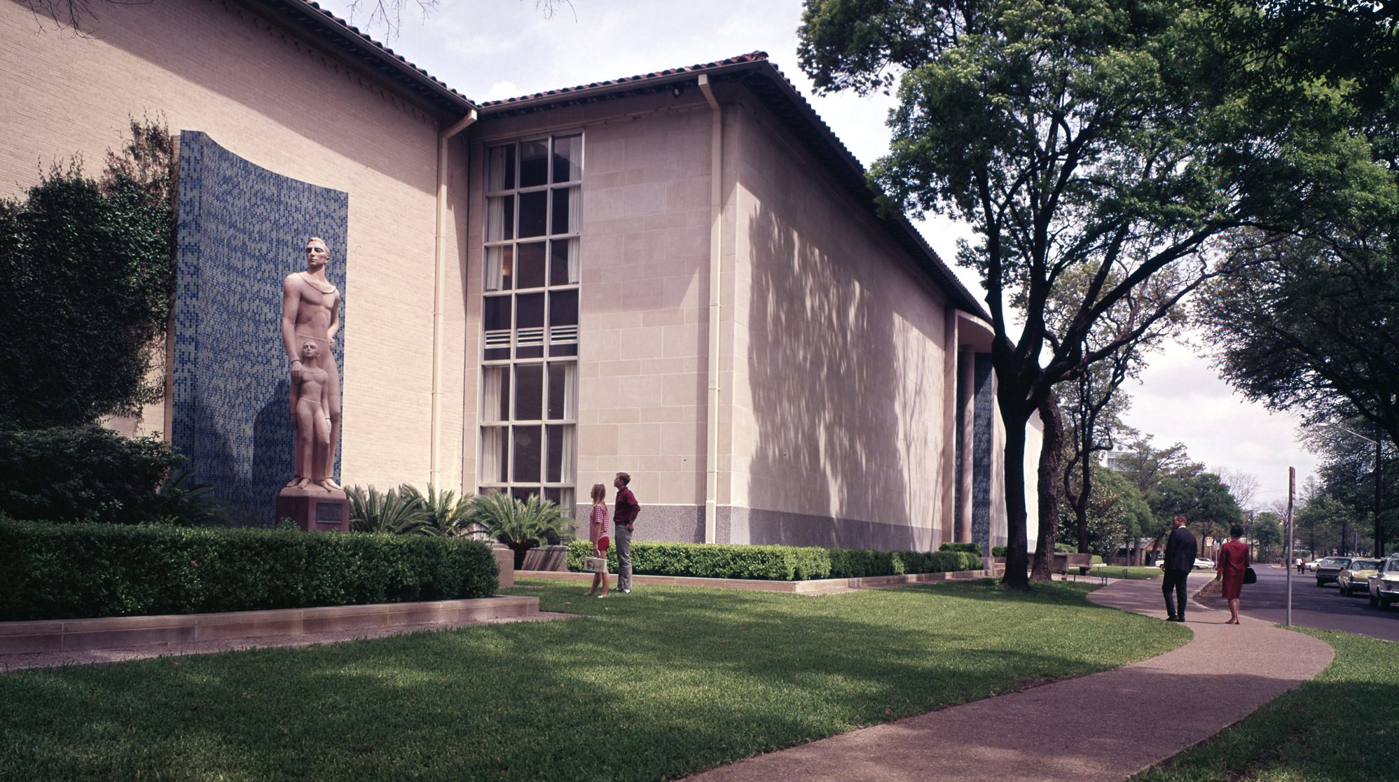 Four people outside a museum, a man and a woman starting at a statue of two figures and the other two people walking down a path lined with trimmed, green grass